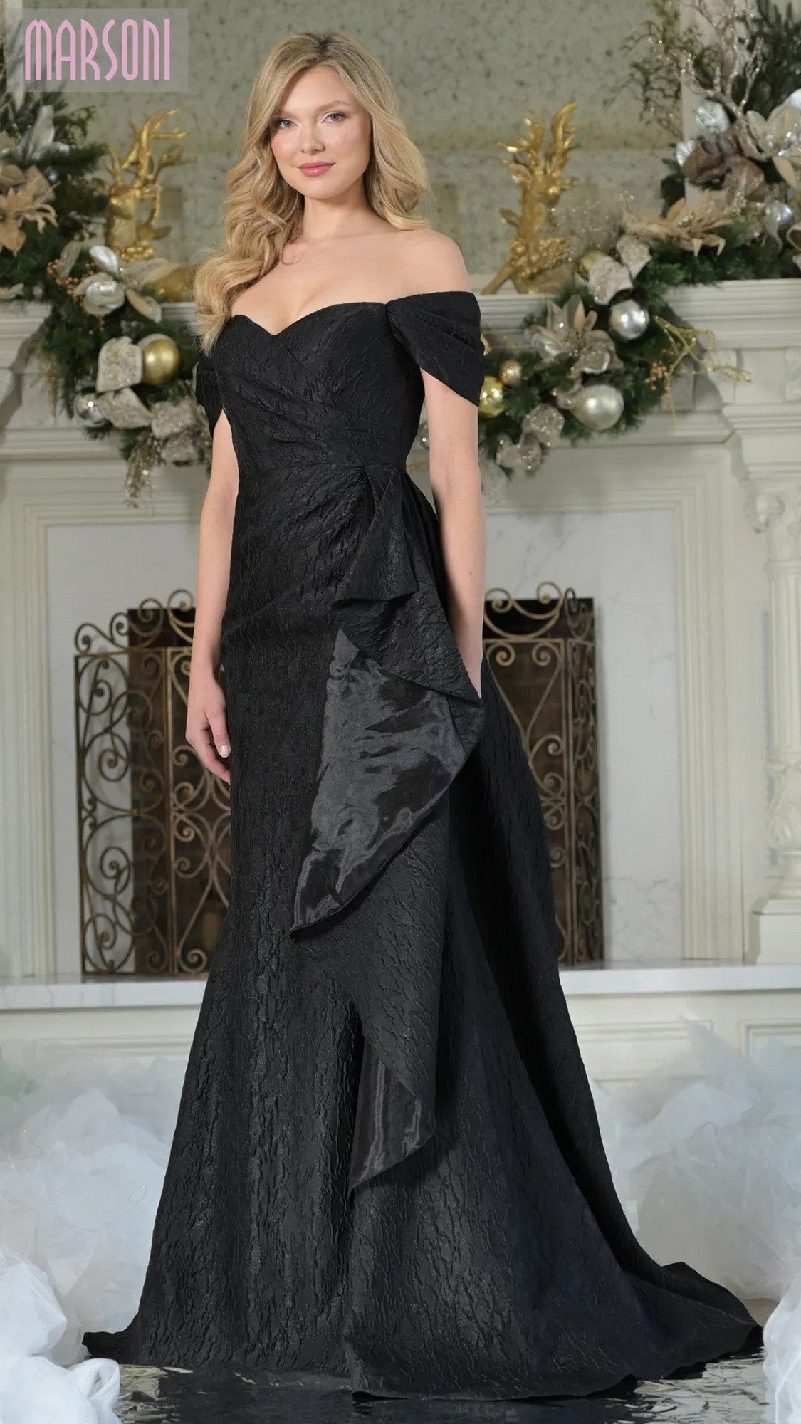A woman stands before a decorated fireplace in the Marsoni MV1365—a black, off-the-shoulder long formal dress featuring textured jacquard fabric, a floor-length sheath silhouette, and a dramatic satin ruffle detail.